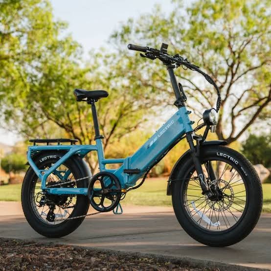 Teal Lectric XP folding step-through e-bike with fat tires, parked on a paved path in a park.