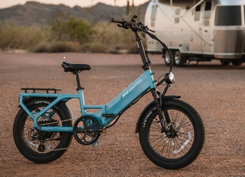 Light blue Lectric folding fat-tire e-bike parked on gravel near an Airstream trailer.
